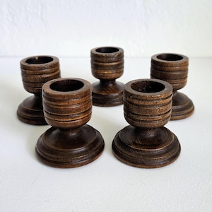 May include: A set of five small, dark brown wooden candle holders. Each holder features a cylindrical top section with three horizontal grooves and a wide, circular base. The wood grain is visible, and the finish is a deep, polished brown tone.