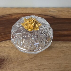 May include: A clear, faceted glass trinket box with a circular lid. The lid features a prominent, textured gold-colored metal flower ornament at its center. The glass body has a geometric, star-like pattern cut into the surface, reflecting light.