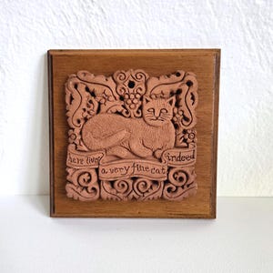 May include: A square wooden plaque featuring a centered terracotta-colored relief carving of a cat resting within ornate scrollwork. A banner across the bottom contains the engraved text: "here lives a very fine cat indeed."