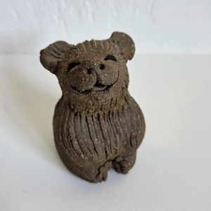 May include: A small, dark brown, textured clay figurine of a bear-like creature. The sculpture features a rounded head with prominent ears, a wide smiling mouth, and a body covered in vertical carved lines that mimic fur, sitting in a seated position.