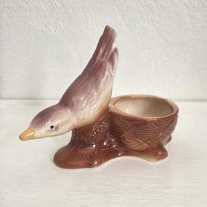 May include: A ceramic figurine featuring a mauve and brown bird perched on the edge of a textured, brown, circular nest-shaped holder. The bird has a yellow beak and a small black eye, while the nest has a woven pattern and a smooth interior.