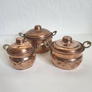 May include: Three copper pots with lids and ornate brass handles. Two smaller pots feature a decorative hammered leaf pattern on the exterior, while the larger central pot has a smooth finish. Each pot includes a matching lid with a central knob.