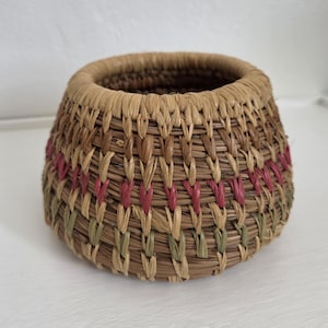 May include: A small, round, hand-woven basket made from natural fibers. The basket features a coiled construction with a thick, smooth rim. The exterior displays a pattern of horizontal bands in muted shades of tan, dark brown, pink, and sage green.