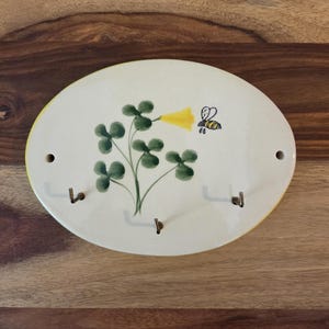 May include: An oval-shaped ceramic wall hook rack with a cream-colored base. The surface features a hand-painted illustration of a green clover plant with a single yellow flower and a small bee. It includes three metal hooks and two mounting holes.