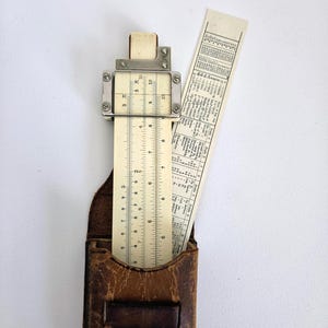 May include: A beige slide rule with a metal cursor and printed numerical scales, partially inserted into a worn brown leather case. The slide rule features a secondary sliding component with printed conversion tables and mathematical data.