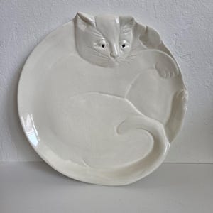 May include: A round, off-white ceramic plate featuring a raised, sculpted relief of a curled-up cat. The cat's face, ears, and tail are integrated into the plate's surface, with small black dots for eyes and etched lines for whiskers.