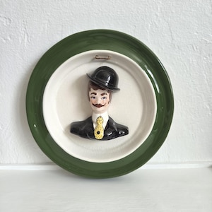 May include: A round ceramic wall plate with a dark green rim and a cream-colored center. A three-dimensional bust of a person wearing a black suit, a white collared shirt, a yellow polka-dot tie, and a black bowler hat is attached to the center.