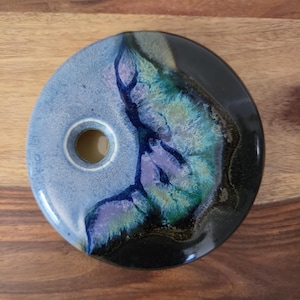 May include: A circular ceramic object with a central hole, featuring a reactive glaze finish. The surface displays a blend of light blue, deep navy, vibrant teal, and purple tones, transitioning into a solid glossy black section on the right side.