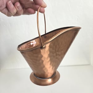May include: A small, hammered copper coal scuttle with a wide, angled opening and a thin, arched metal handle. The vessel features a polished, metallic finish with a textured, dimpled surface and a circular base for stability.