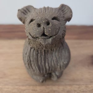 May include: A small, dark gray, textured ceramic figurine of a bear with a smiling face, rounded ears, and vertical carved lines on its body. The sculpture has a rough, earthy finish and is depicted in a seated position with a cheerful expression.