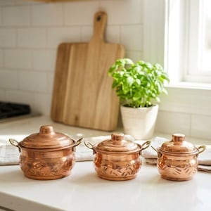 May include: Three round copper pots with lids and side handles, arranged in descending order of size. Each pot features a polished metallic finish and a textured, embossed pattern around the lower body, showcasing a warm, reflective copper tone.