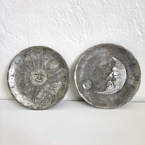 May include: Two circular metal plates with a dark, weathered finish. One plate features a raised sun face with radiating rays, while the other displays a crescent moon face with a small circular orb, both surrounded by tiny star accents.