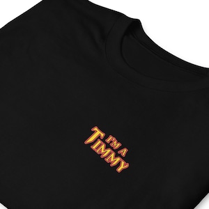 May include: Black crew neck t-shirt with the embroidered text "I'm a Timmy" in yellow and red. The text is in a stylized font. The shirt is made of a soft material.
