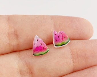 Könnte beinhalten: Ein Paar Ohrstecker in Form kleiner Wassermelonenstücke. Jeder Ohrstecker zeigt ein leuchtend rosa Fruchtfleisch, eine dünne weiße Schale und einen grünen Außenrand, mit kleinen schwarzen, tropfenförmigen Kernen auf der rosa Oberfläche.