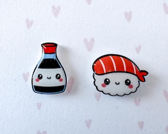 Könnte beinhalten: Zwei dekorative Anstecknadeln mit Sushi- und Sojasaucenflaschen-Motiven im Cartoon-Stil. Die Sojasaucenflasche ist schwarz, weiß und rot mit einem lächelnden Gesicht. Das Sushi-Stück zeigt eine weiße Reisbasis mit einem rot-weiß gestreiften Fischstück.