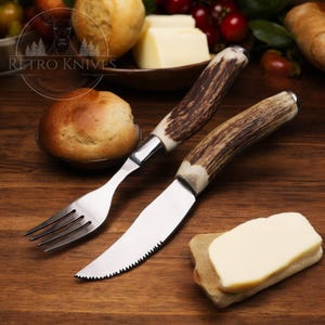 May include: A set of two kitchen knives with antler handles. The knife on the left is a fork and the knife on the right is a serrated butter knife. The knives are on a wooden surface with a piece of bread and butter.