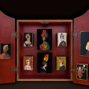 May include: A red wooden cabinet with a hinged door, revealing a series of small framed portraits of people wearing elaborate hats. The portraits are arranged in a grid pattern and are set against a gold background.