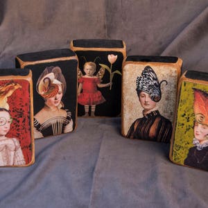 May include: Five wooden blocks, each featuring a different image of a woman wearing a unique hat. The blocks have a black background and each image depicts a woman with a distinctive hat made from feathers, butterflies, and other materials.