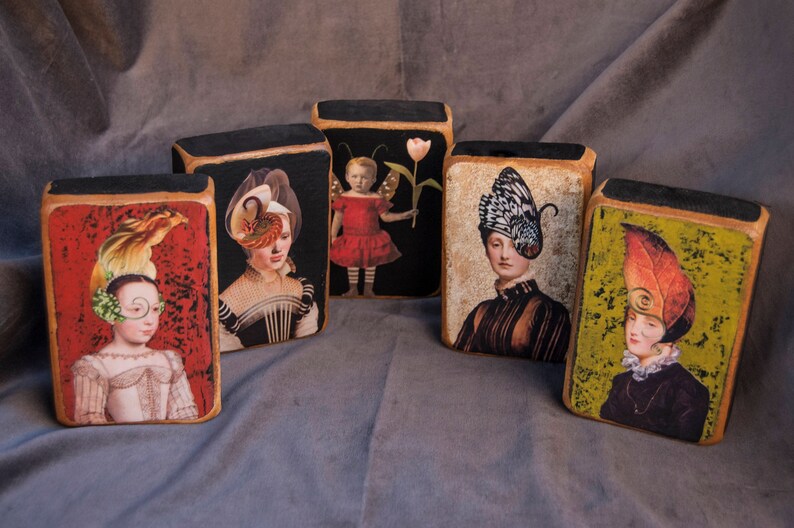 May include: Five wooden blocks, each featuring a different image of a woman wearing a unique hat. The blocks have a black background and each image depicts a woman with a distinctive hat made from feathers, butterflies, and other materials.