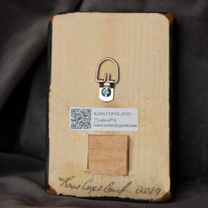 May include: The back of a wooden framed painting with a QR code, contact information, and the artist's signature. The signature reads "Kass Copeland 2017".