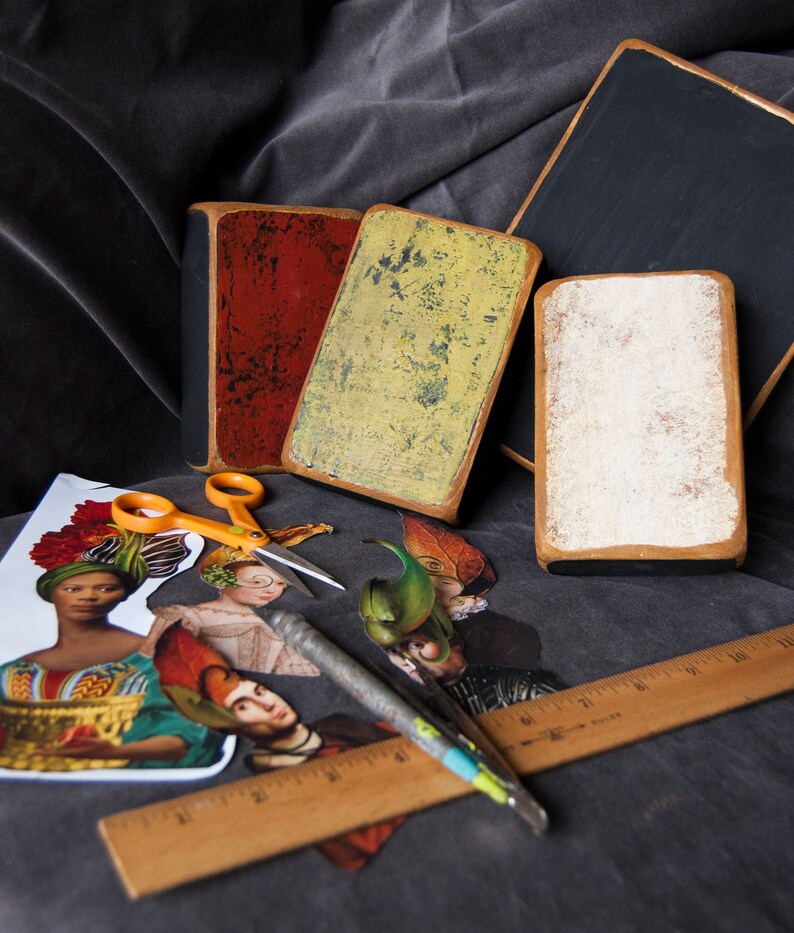 May include: Four vintage-style wooden signs with distressed paint finishes. The signs are in various colours, including red, yellow, black, and white. The signs are arranged on a dark fabric surface with a ruler, scissors, and craft paper.
