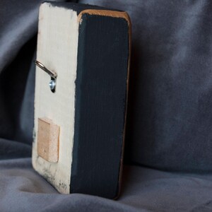 May include: A small wooden sign with a white background and black painted edges. The sign has a metal hook on the top and a small wooden rectangle attached to the back. The sign is sitting on a grey fabric surface.