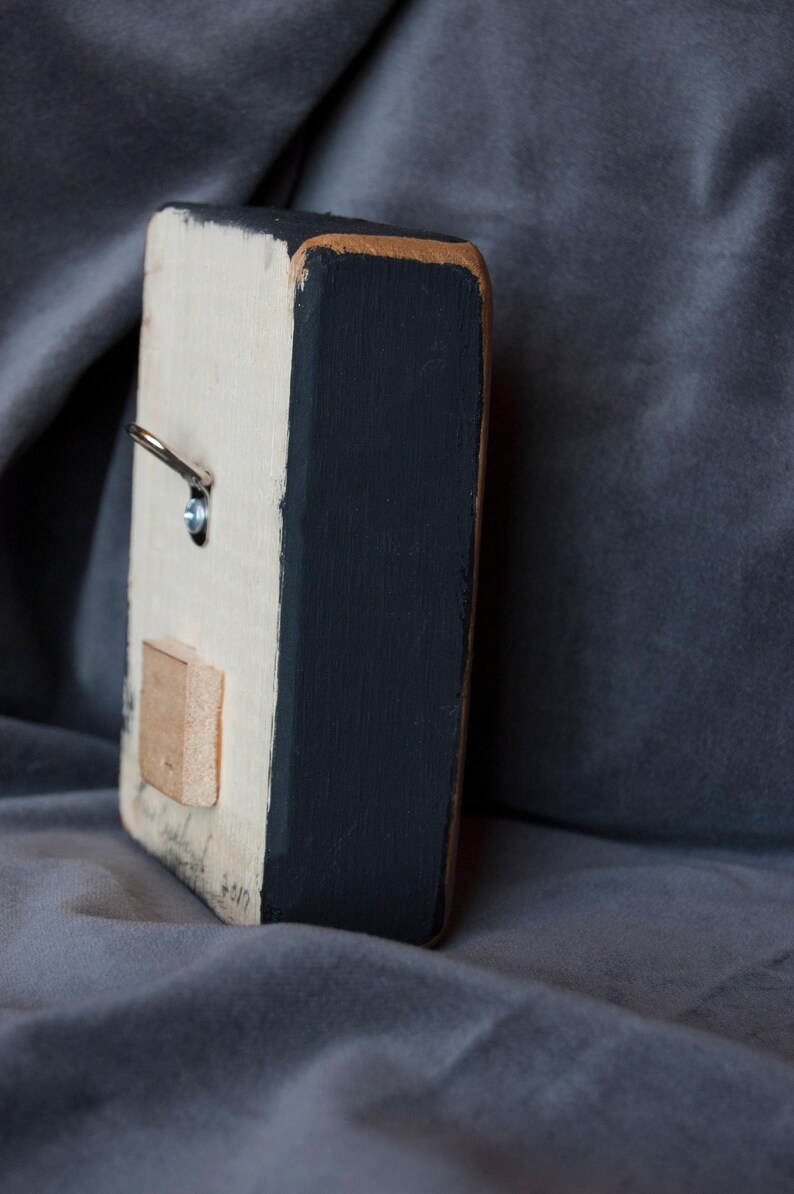 May include: A small wooden sign with a white background and black painted edges. The sign has a metal hook on the top and a small wooden rectangle attached to the back. The sign is sitting on a grey fabric surface.