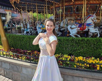May include: A woman in a white iridescent dress with a gold belt stands in front of a carousel. She is smiling and looking at the camera. The carousel is decorated with colorful horses and lights.