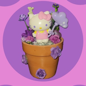 Könnte beinhalten: Ein Terrakotta-Blumentopf, verziert mit lila Blumen und einer Hello Kitty-Figur, die einen lila Ballon hält. Der Topf steht auf einem lila Hintergrund.