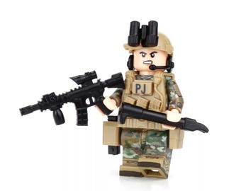 May include: A camouflage-patterned Lego minifigure of a soldier wearing a helmet with a black visor and a black headset. The figure is holding a black rifle and a black pistol. The figure has a tan vest with the letters "PJ" printed on it.