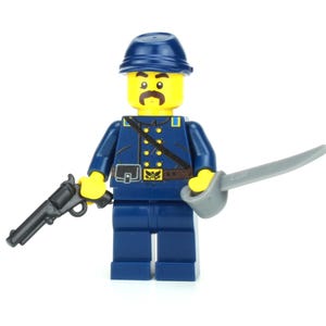 May include: A blue and yellow Lego minifigure of a soldier wearing a blue hat, a blue jacket, and a brown belt. The figure is holding a black pistol in one hand and a silver sword in the other.