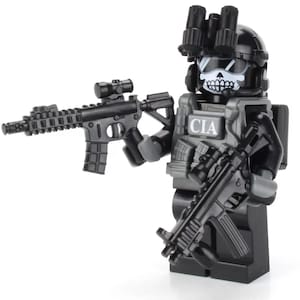 May include: A black and grey Lego minifigure of a soldier wearing a black helmet with a skull face mask and a black vest with the letters "CIA" printed on it. The minifigure is holding two black guns.