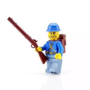 May include: A blue and yellow Lego minifigure of a Union soldier holding a brown rifle. The soldier is wearing a blue uniform with a yellow belt and a blue hat. The figure has a backpack and the text "US" on the belt.