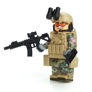 May include: A toy soldier minifigure wearing a camouflage uniform, a tan helmet, and goggles. The figure is holding a black toy rifle with a scope. The figure is wearing a tan tactical vest with pouches.