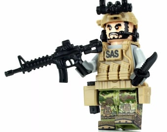 May include: A Lego minifigure of a soldier wearing a tan camouflage uniform, a black helmet with a red dot sight, and a black vest with the word "SAS" on it. The soldier is holding a black rifle and a black knife.
