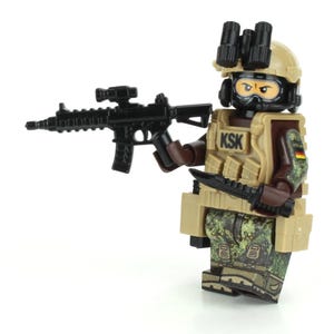 Puede incluir: Una minifigura de Lego de un soldado alemán con uniforme de camuflaje, un casco negro con visor de visión nocturna y un chaleco negro con el texto "KSK". La figura sostiene un rifle negro y un cuchillo negro.