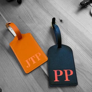 May include: Two leather luggage tags with initials. The tag on the left is orange with the initials "JTP" in pink. The tag on the right is black with the initials "PP" in pink.