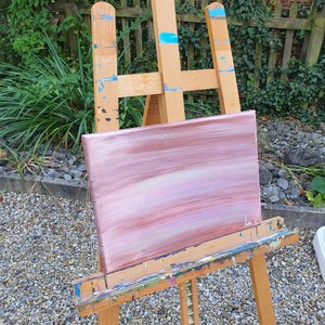 May include: A pink and white abstract painting on a wooden easel. The painting has horizontal brushstrokes and is set against a background of small grey stones.