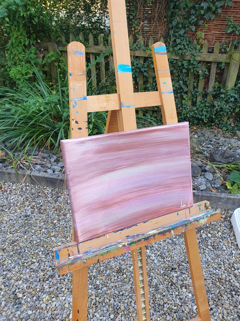 May include: A pink and white abstract painting on a wooden easel. The painting has horizontal brushstrokes and is set against a background of small grey stones.
