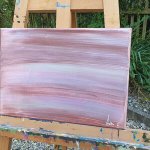 May include: Abstract painting on canvas with horizontal brushstrokes in shades of pink, white, and brown. The painting is on an easel.
