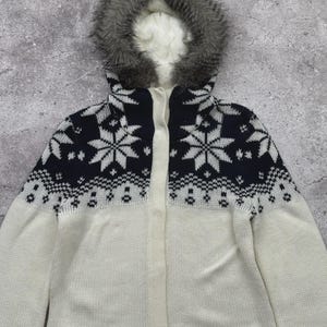 May include: A white and navy blue knit hooded jacket with a snowflake pattern. The jacket features a faux fur-lined hood and a full-length zipper. The sleeves and lower body are white, while the upper chest area is navy blue with white snowflake designs.