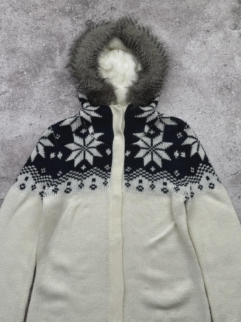 May include: A white and navy blue knit hooded jacket with a snowflake pattern. The jacket features a faux fur-lined hood and a full-length zipper. The sleeves and lower body are white, while the upper chest area is navy blue with white snowflake designs.