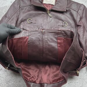 K&ouml;nnte beinhalten: Burgunderfarbene Lederjacke mit Rei&szlig;verschluss, Kragen und zwei Vordertaschen. Die Jacke hat ein Satinfutter und goldfarbene Hardware. Der Markenname "Vanguard" ist auf einem Etikett im Inneren sichtbar.