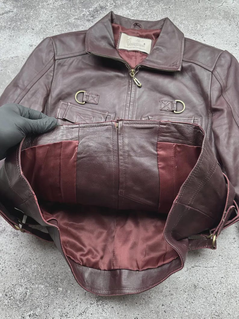 K&ouml;nnte beinhalten: Burgunderfarbene Lederjacke mit Rei&szlig;verschluss, Kragen und zwei Vordertaschen. Die Jacke hat ein Satinfutter und goldfarbene Hardware. Der Markenname "Vanguard" ist auf einem Etikett im Inneren sichtbar.