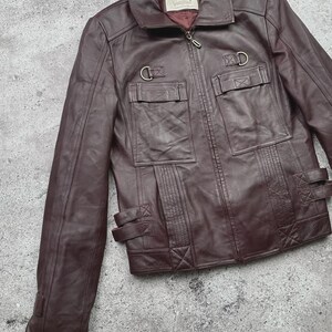 K&ouml;nnte beinhalten: Burgunderfarbene Lederjacke mit Rei&szlig;verschluss, zwei Brusttaschen und zwei unteren Taschen. Die Jacke hat einen Kragen und dekorative Metallringe und Riemen. Die Jacke ist aus glattem, geschmeidigem Leder gefertigt.