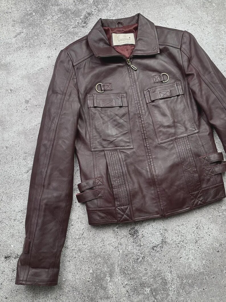 K&ouml;nnte beinhalten: Burgunderfarbene Lederjacke mit Rei&szlig;verschluss, zwei Brusttaschen und zwei unteren Taschen. Die Jacke hat einen Kragen und dekorative Metallringe und Riemen. Die Jacke ist aus glattem, geschmeidigem Leder gefertigt.