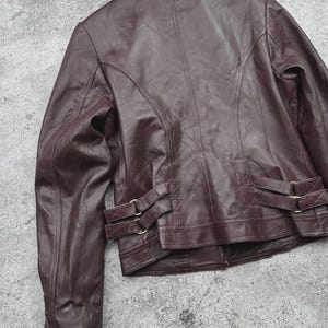 K&ouml;nnte beinhalten: Burgunderfarbene Lederjacke mit klassischem Kragen und taillierter Passform. Die Jacke hat dekorative N&auml;hte, einen Knopfverschluss und verstellbare Riemen mit goldfarbenen Schnallen an der Taille. Das Leder hat ein leichtes Used-Aussehen.