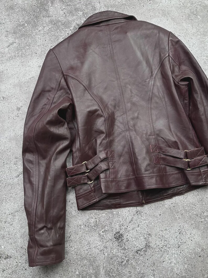 K&ouml;nnte beinhalten: Burgunderfarbene Lederjacke mit klassischem Kragen und taillierter Passform. Die Jacke hat dekorative N&auml;hte, einen Knopfverschluss und verstellbare Riemen mit goldfarbenen Schnallen an der Taille. Das Leder hat ein leichtes Used-Aussehen.