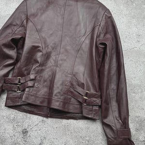 K&ouml;nnte beinhalten: Burgunderfarbene Lederjacke mit klassischem Design. Die Jacke hat einen Reverskragen, lange &Auml;rmel und dekorative Schnallen an der Taille. Die R&uuml;ckseite der Jacke hat vertikale N&auml;hte f&uuml;r eine taillierte Passform. Das Leder hat eine glatte, leicht gl&auml;nzende Oberfl&auml;che.