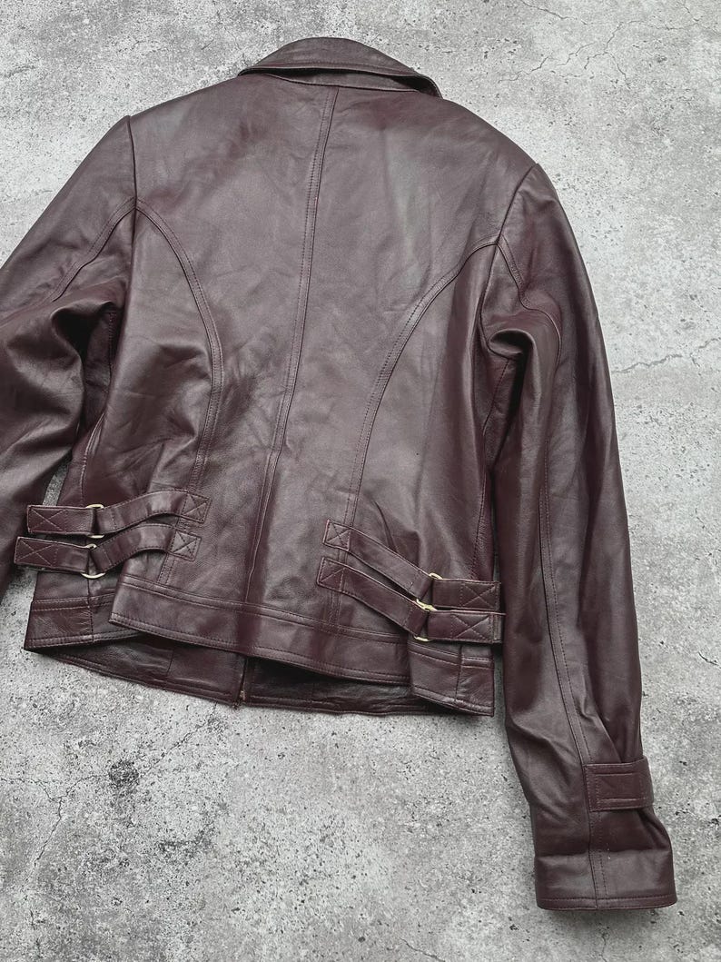 K&ouml;nnte beinhalten: Burgunderfarbene Lederjacke mit klassischem Design. Die Jacke hat einen Reverskragen, lange &Auml;rmel und dekorative Schnallen an der Taille. Die R&uuml;ckseite der Jacke hat vertikale N&auml;hte f&uuml;r eine taillierte Passform. Das Leder hat eine glatte, leicht gl&auml;nzende Oberfl&auml;che.